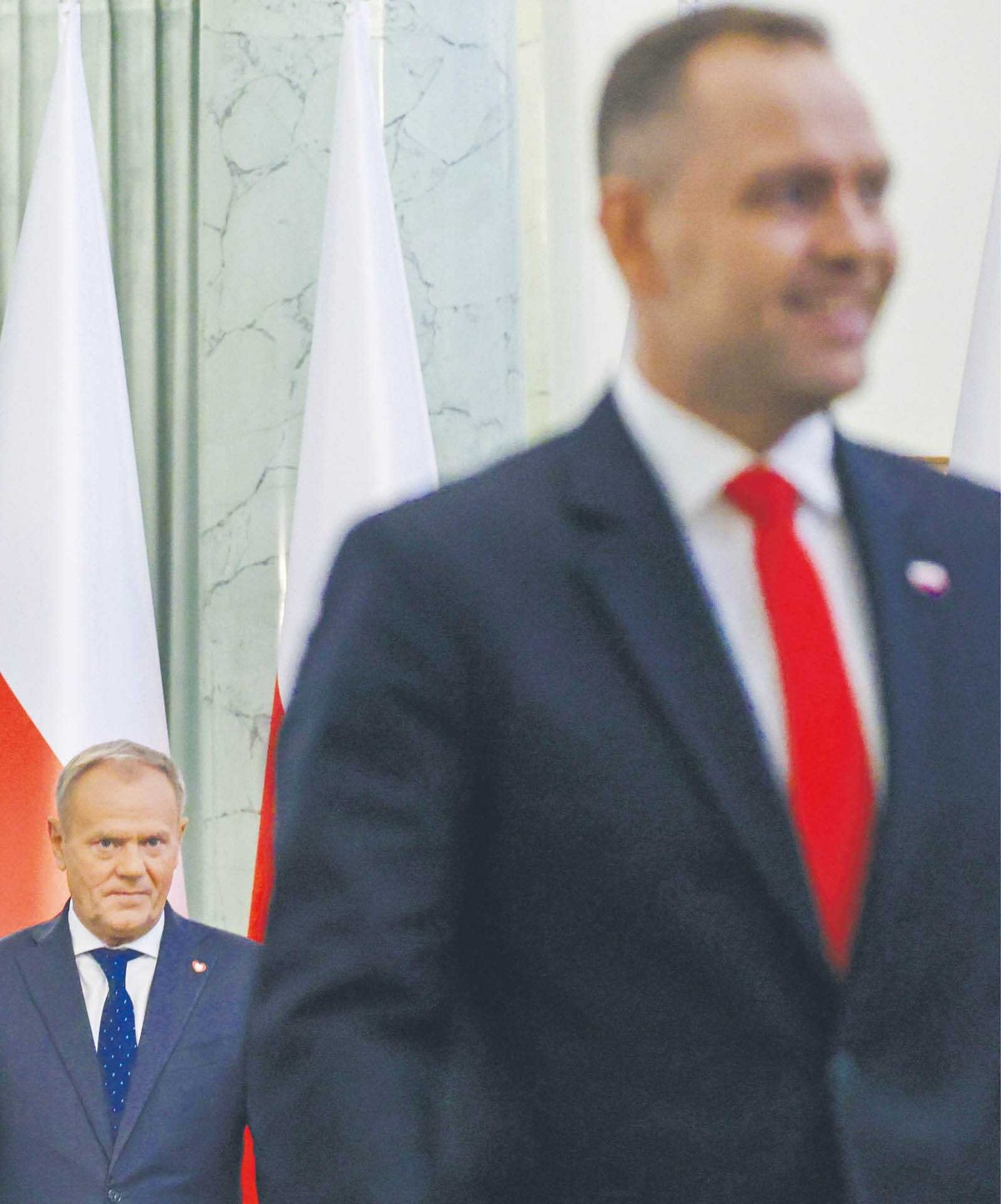 Na pierwszym planie widoczny jest uśmiechnięty prezydent RP Karol Nawrocki w garniturze z czerwonym krawatem. W tle, na tle polskich flag, widać premiera Donalda Tuska z poważnym wyrazem twarzy