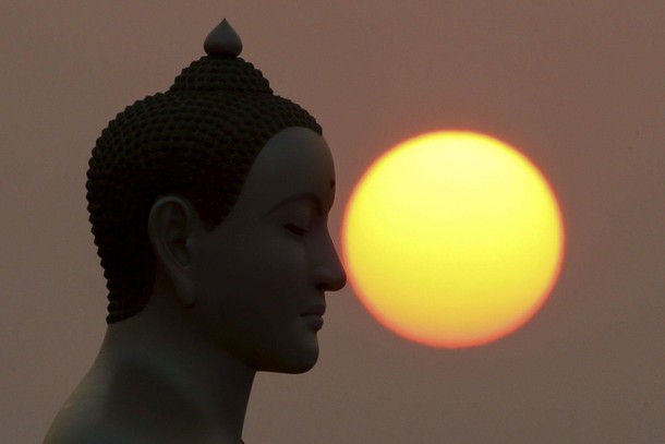 The sun rises next to a Buddha statue while monks and novices gather to receive alms at Wat Phra Dha