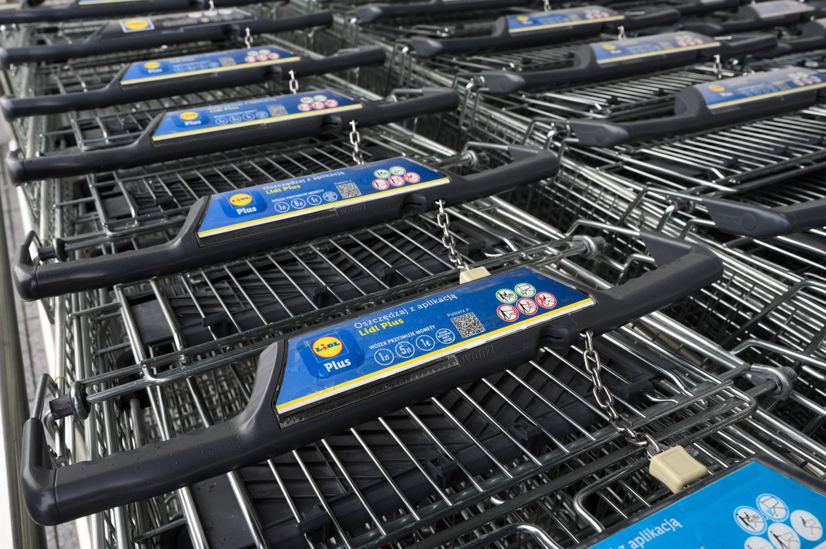 Rząd zakupowych wózków sklepowych przed sklepem Lidl, z widocznymi uchwytami i informacją o aplikacji Lidl Plus.