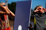 A reflected image of the sun is seen on a white board as kids look up to view the beginning a partia