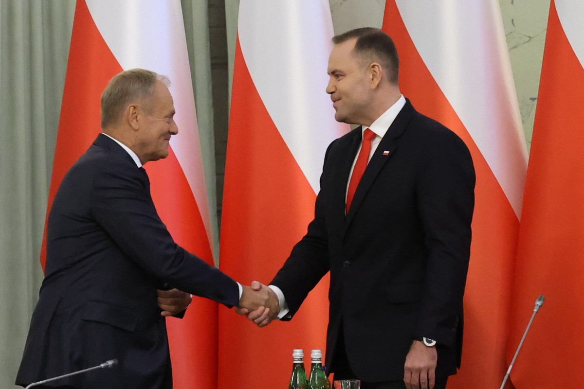 Premier Donald Tusk (po lewej) i prezydent Karol Nawrocki (po prawej) podają sobie ręce podczas spotkania, w tle widoczne biało-czerwone flagi Polski.