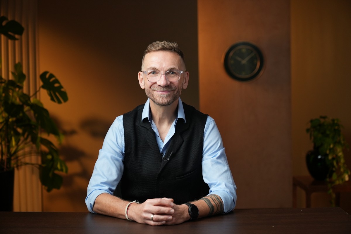 Przemek Kossakowski, dziennikarz i ambasador kampanii WSKZ ‚Kierunek: edukacja’, siedzi przy biurku, uśmiecha się, trzymając złożone ręce, w tle zegar i kwiatek doniczkowy