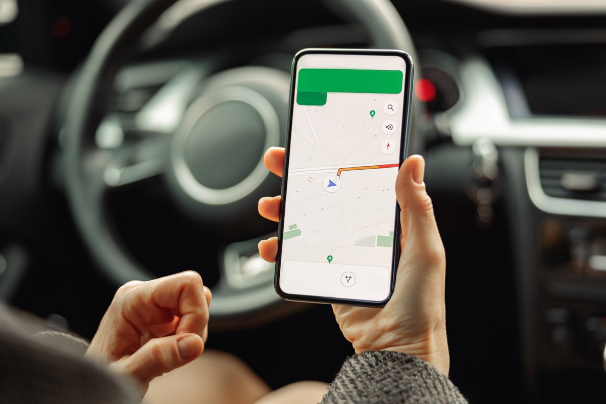 Osoba siedząca w samochodzie trzyma smartfon z włączoną aplikacją nawigacyjną pokazującą trasę.