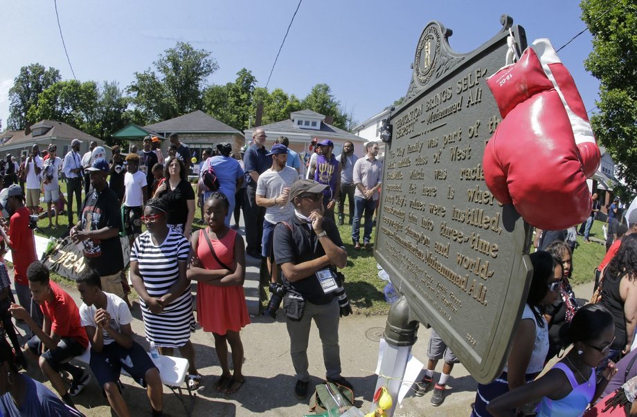 Rękawice bokserskie przewieszone na płycie pamiątkowej przed domem rodzinnym Muhammada Alego. Ludzie oczekują na przyjazd konduktu pogrzebowego. 