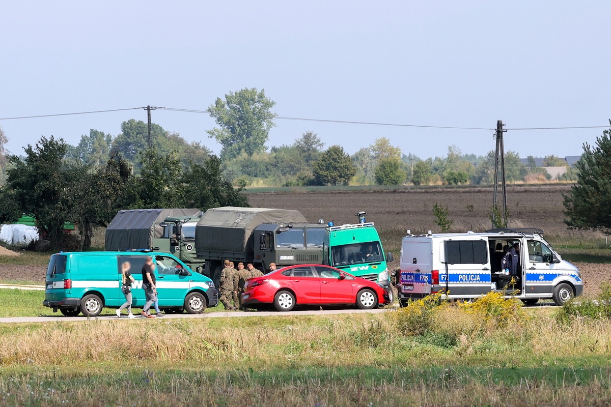 Wojsko i policja przy pojazdach na wiejskiej drodze, w tle pola i drzewa – akcja służb w terenie.