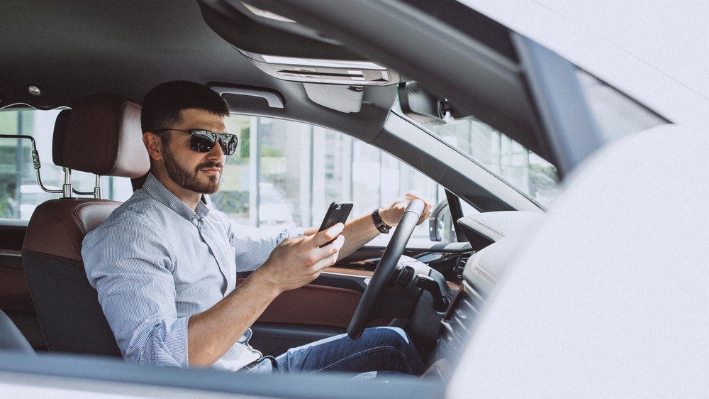 Mobilný telefón v ruke vodiča na parkovisku pred autoservisom, na obrazovke otvorená aplikácia Google Mapy s profilom dielne s priemerom 4,9 hviezdičky a tristo recenziami, vodič palcom posúva nadol zoznam hodnotení, kde vidno tri zhodne formulované päťhviezdičkové komentáre po sebe – ilustrácia typického vzorca kúpených fake recenzií na slovenskom trhu autoservisov. 