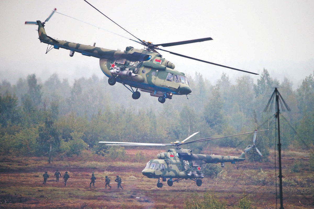 Na zdjęciu widać dwa wojskowe śmigłowce – prawdopodobnie rosyjskie Mi-8 lub Mi-17 – wykonujące manewry nad otwartym terenem, najpewniej podczas ćwiczeń wojskowych. W tle widoczna jest grupa żołnierzy oraz zalesiony obszar, co sugeruje działania taktyczne w terenie przygranicznym lub poligonowym. Scena pokazuje intensywny charakter działań zbrojnych i przygotowanie do operacji powietrzno-lądowych