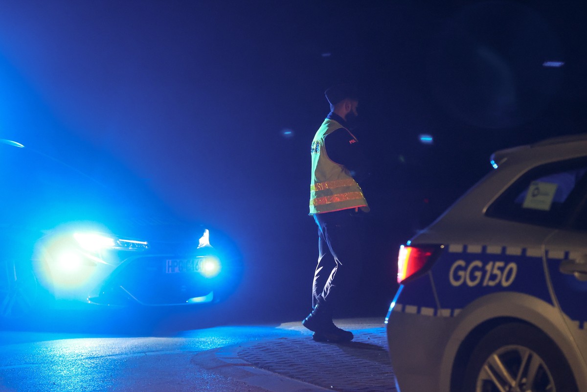 Policjant w odblaskowej kamizelce stoi nocą na drodze, obok radiowozów z włączonymi niebieskimi światłami sygnalizacyjnymi, które oświetlają okolicę intensywną, niebieską poświatą.