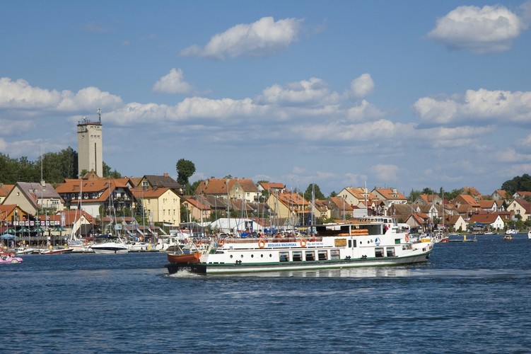 <strong>Mikołajki</strong>
<br></br>
Mikołajki to niewielkie miasteczko oblane Jeziorem Mikołajskim i Tałty. To port docelowy wielu żeglarskich rejsów po mazurskich wodach. Mekka dla wodniaków, czyli amatorów rekreacji w akwenach. Dla tych którzy nie odważą się na popłynięcie żaglówką czekają tu np. kajaki. Kiedy już nasycimy wzrok przyrodą widzianą znad kajakowego wiosła - warto wejść także na ląd i zobaczyć staromiejską, zabytkową zabudowę, gdzie widoczne są m.in wpływy reformacji.