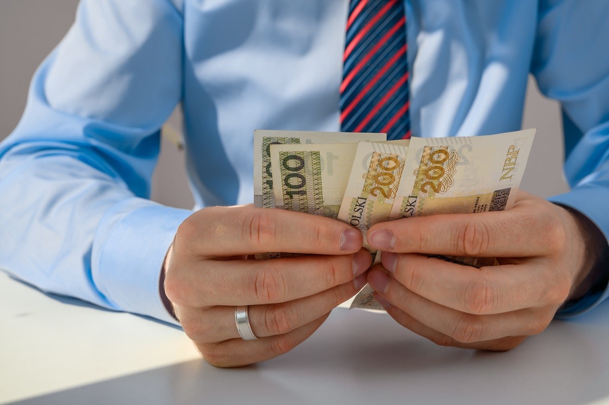 Mężczyzna w koszuli i krawacie trzyma w dłoniach banknoty polskich złotych.