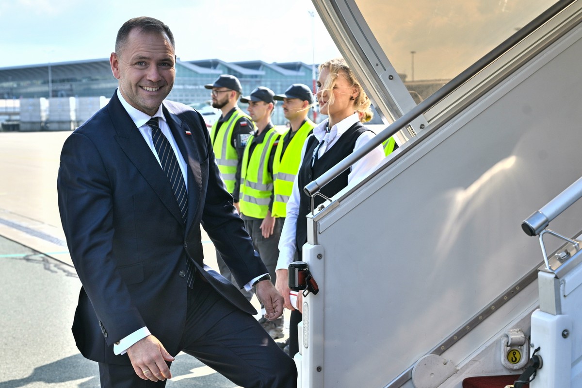 Prezydent Polski Karol Nawrocki w garniturze wchodzi po trapie samolotu, uśmiechając się do kamery. W tle stoją pracownicy lotniska w kamizelkach odblaskowych.