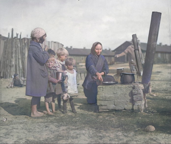 Grupa dzieci na dworze przy prowizorycznej kuchni, na której kobieta przyrządza posiłek. 1934 rok. Źródło: Narodowe Archiwum Cyfrowe.
