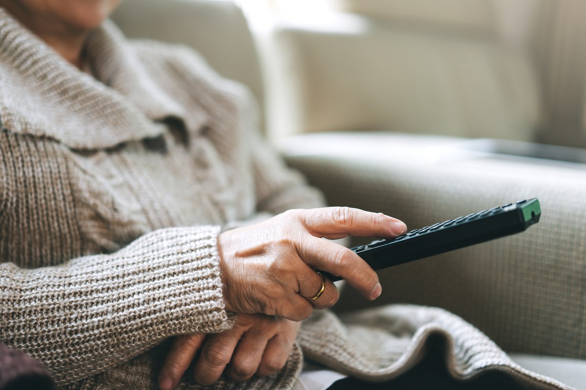 Starsza osoba w szarej dzianinowej bluzie siedzi na kanapie i trzyma w dłoni pilot do telewizora.