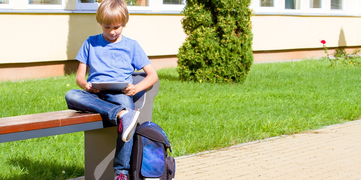 Schoolboy with tablet PC sitting on the bench near school. Outdoor. Education, technology, people co