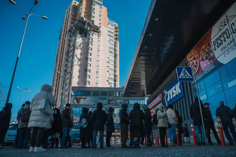 Mieszkańcy Kijowa stoją w kolejce do jednego z nielicznych otwartych supermarketów. W tle uszkodzony przez rakietę blok mieszkalny (28 lutego2022 r., Kijów) 