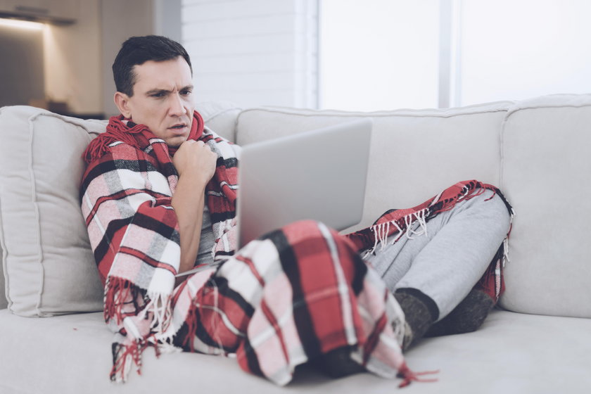A man with a cold sits on the couch, hiding behind a red rug. He is sitting with his laptop on his l