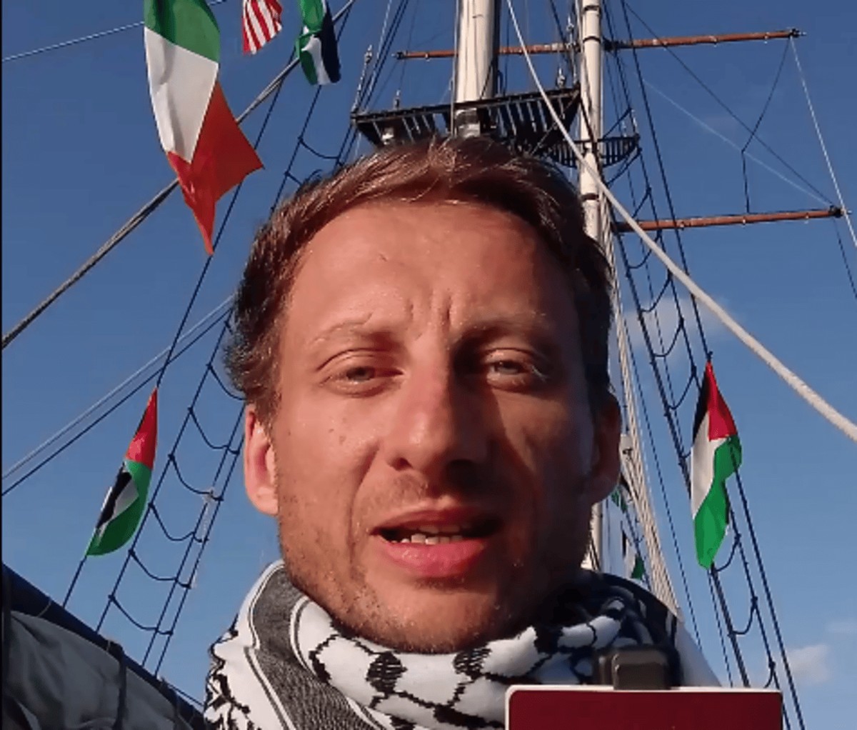 Starczewski w chustce typu kefija mówi do kamery na tle masztu statku z powiewającymi flagami, w tym palestyńskimi, irlandzką i amerykańską.