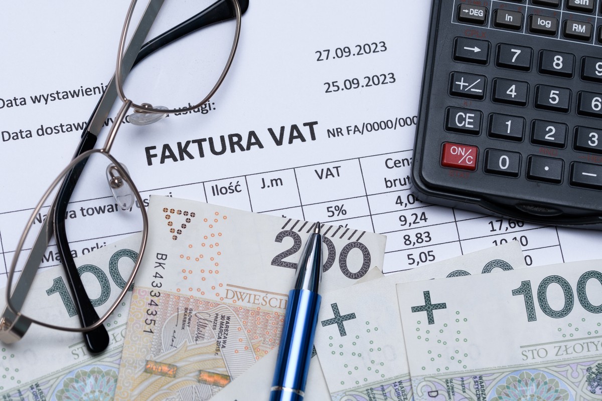 Dokument faktury VAT z datami, polskimi banknotami, długopisem, okularami i kalkulatorem na biurku. Przedstawienie tematyki księgowości, podatku VAT, finansów i KSeF.