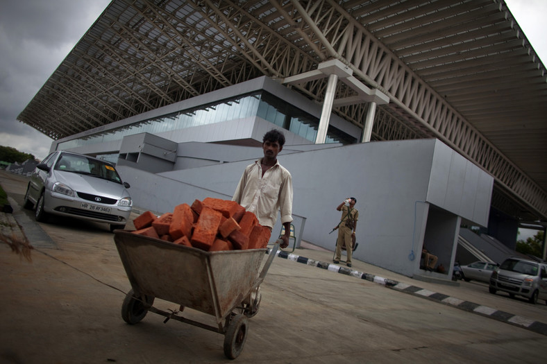 Robotnik budowlany pcha taczki pełne cegieł przed prawie gotową halą Thyagaraj Sport Complex w New Delhi w Indiach. Na tym obiekcie będą rozgrywane mecze koszykówki podczas Igrzysk Wspólnoty Narodów 2010. Fot.: Brian Sokol/Bloomberg