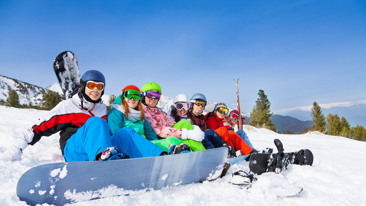 Grupka przyjaciół siedzi na śniegu z deskami snowboardowymi i nartami na tle gór