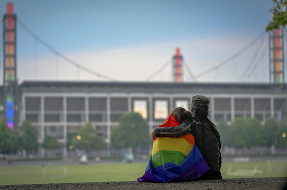 Osoby owinięte tęczową flagą obejmują się przed oświetlonym stadionem RheinEnergieStadion w Kolonii