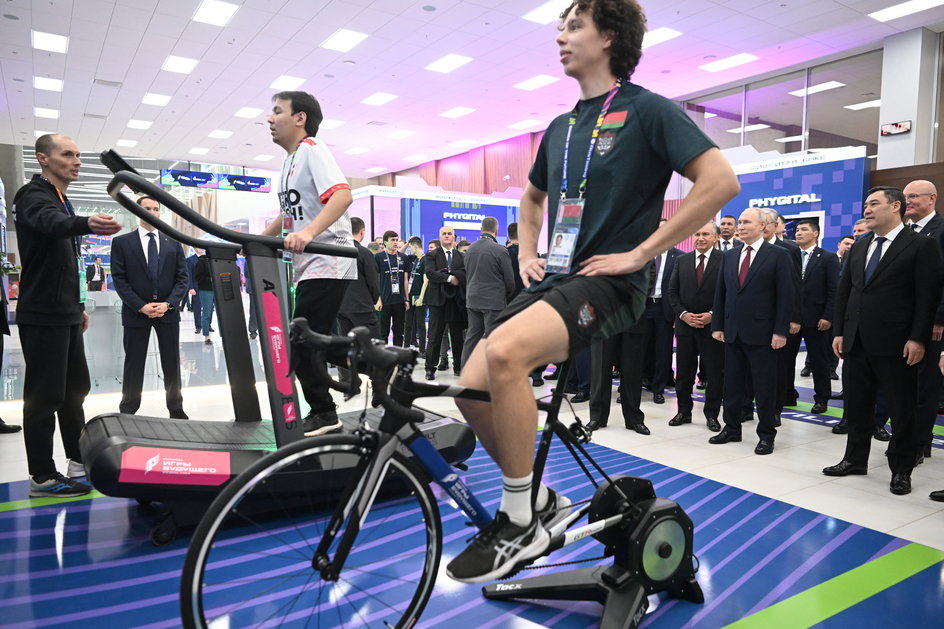 Trenażer rowerowy na Igrzyskach Przyszłości. W tle Władimir Putin